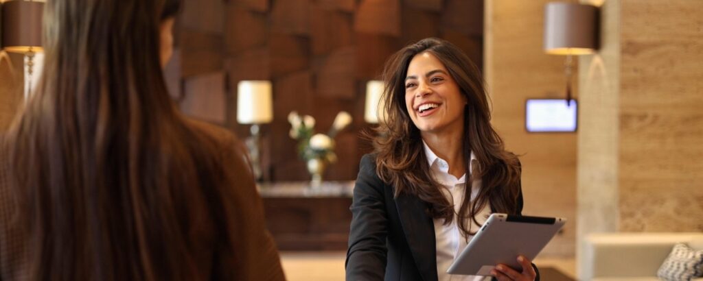 Header für den Beitrag ‚Mitarbeitergespräch erfolgreich führen: So gelingt es‘. Darauf eine lächelnde Frau im Business-Outfit mit Tablet, die jemandem die Hand gibt.