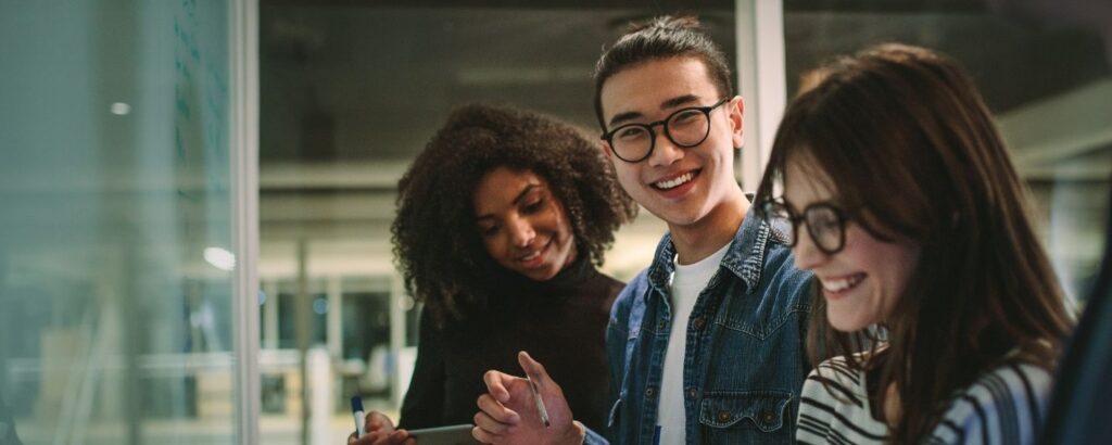 Ratgeber 6 Drei junge Menschen arbeiten gemeinsam an einem Projekt in einem modernen Büro. Eine Person im Vordergrund lächelt in die Kamera, während die anderen konzentriert Notizen machen – Symbol für Teamarbeit, Motivation und den Einstieg als Junior Consultant.