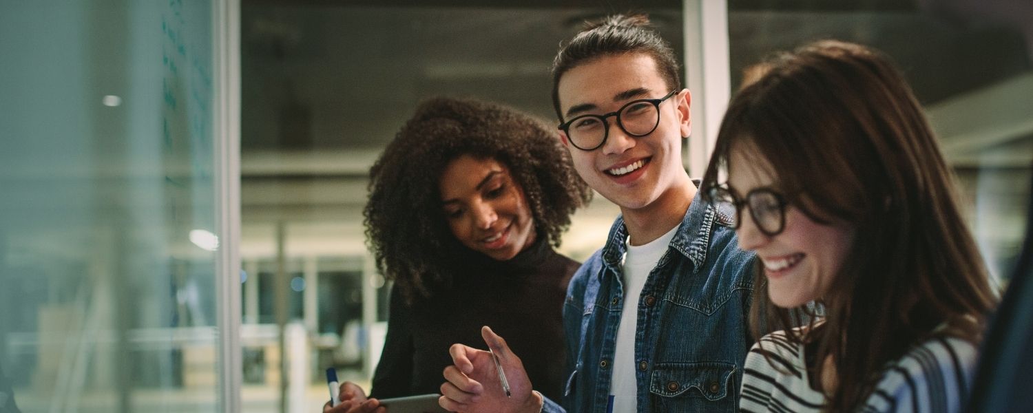 Drei junge Menschen arbeiten gemeinsam an einem Projekt in einem modernen Büro. Eine Person im Vordergrund lächelt in die Kamera, während die anderen konzentriert Notizen machen – Symbol für Teamarbeit, Motivation und den Einstieg als Junior Consultant.