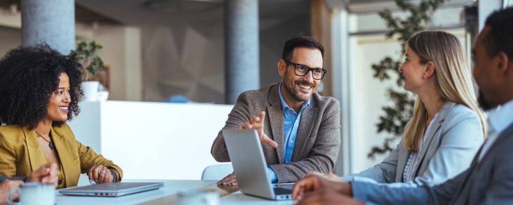 Mehrere berufstätige Personen sitzen in einem Meetingraum zusammen, tragen Business-Casual-Outfits und führen ein freundliches Gespräch. Ein Mann mit Brille gestikuliert lächelnd, während die anderen aufmerksam zuhören.