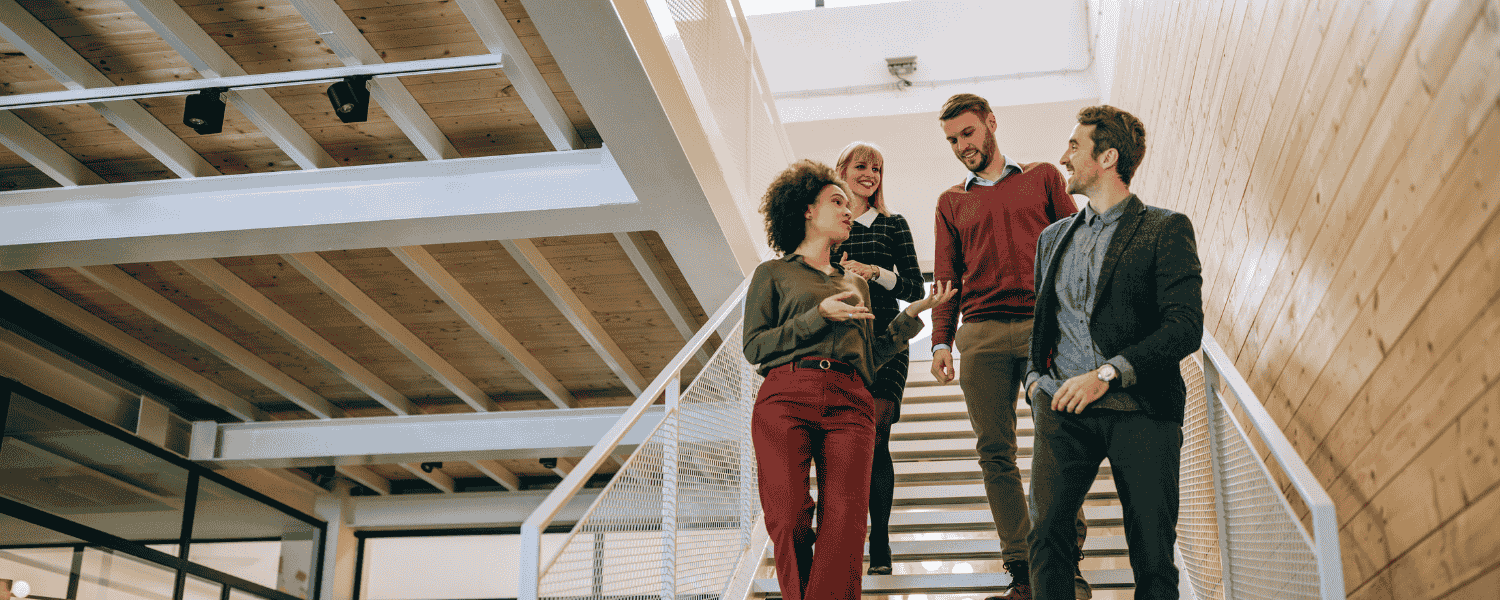 Vier Kolleginnen und Kollegen in Business-Casual-Kleidung gehen eine moderne Bürotreppe hinunter und unterhalten sich in einer offenen, hellen Arbeitsumgebung.