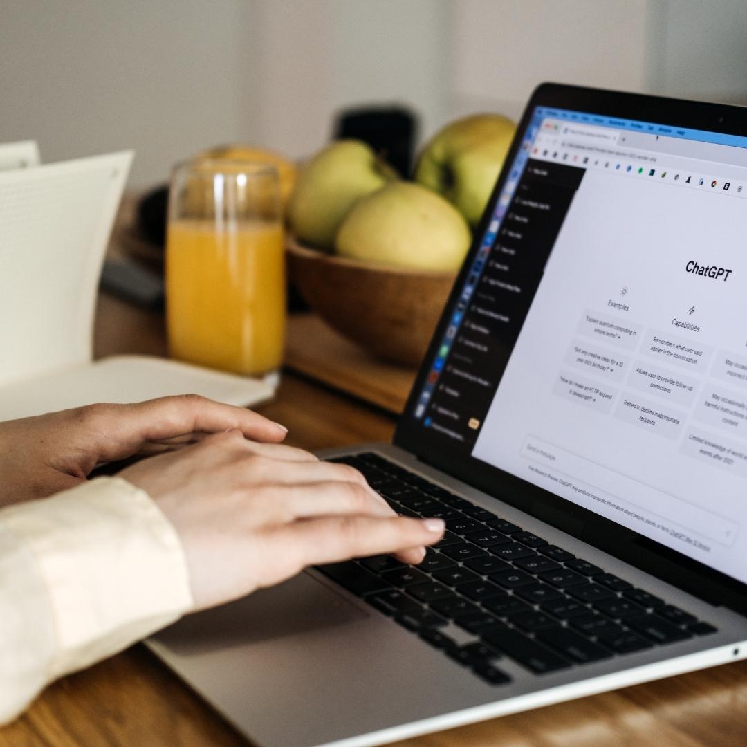 Person tippt an einem Laptop mit geöffneter ChatGPT-Oberfläche auf dem Bildschirm, auf dem Schreibtisch stehen ein Glas Orangensaft und eine Schale mit Äpfeln.