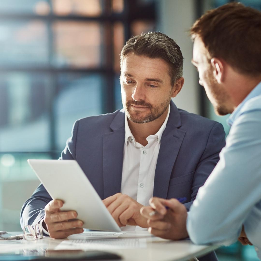 Zwei Personen sitzen an einem Tisch und führen eine Verhandlung. Eine Person hält ein Tablet in der Hand.