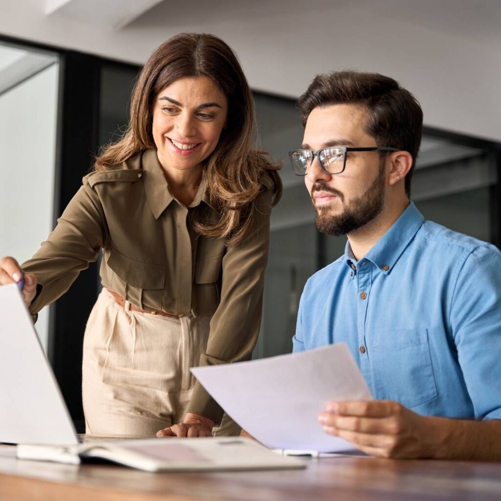Zwei Personen arbeiten gemeinsam in einem Büro: Ein Mann im Hemd sitzt vor einem Laptop, während eine Frau in Bluse danebensteht und auf den Bildschirm zeigt; beide schauen konzentriert auf den Laptop.