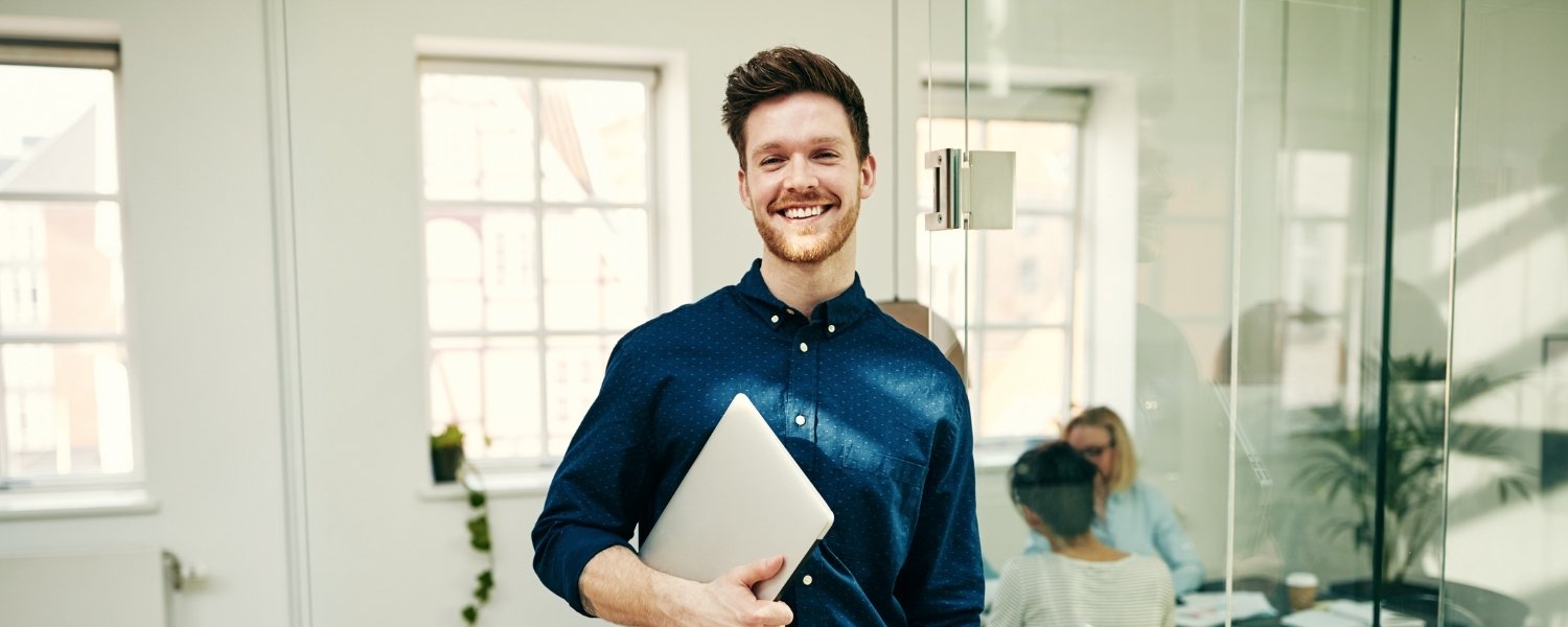 Ein junger Mann lächelt in die Kamera und hält einen Laptop in den Händen. Er steht in einem hellen Büro mit Glaswänden, im Hintergrund arbeiten weitere Personen an einem Tisch.