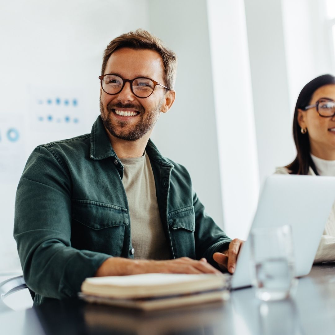 Ein Mann sitzt an einem Schreibtisch, arbeitet am Laptop und lächelt. Neben ihm arbeitet eine Frau.