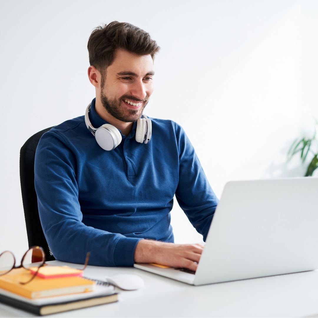 Junger Mann sitzt am Schreibtisch vor dem Laptop, trägt Kopfhörer um den Hals, lächelt und arbeitet konzentriert.