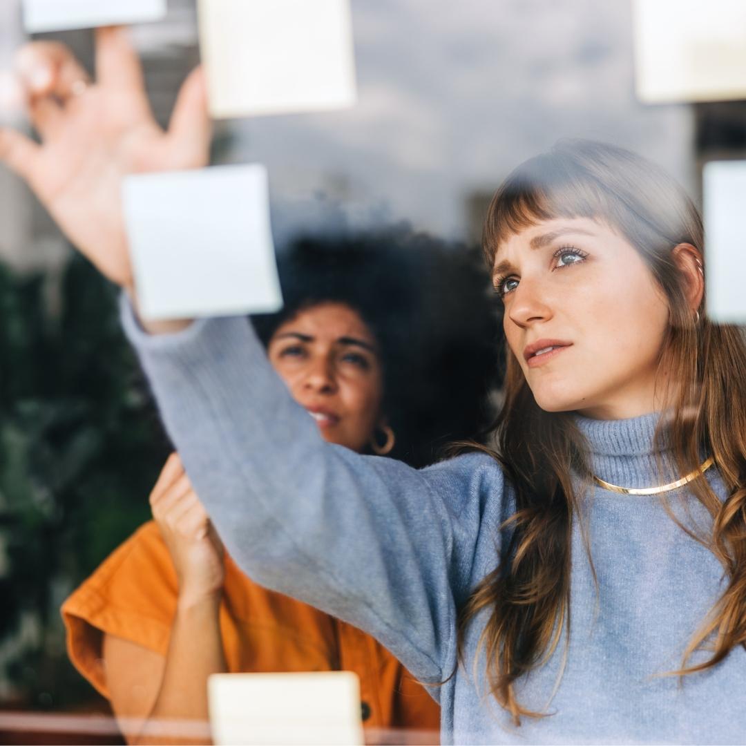 Zwei Frauen arbeiten im Büro an einem durchsichtigen Whiteboard mit Post-its und planen gemeinsam Projekte. Symbolisch für Hands on Mentalität.