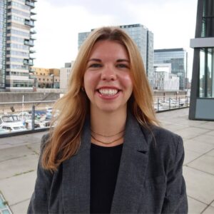 Porträt von Johanna Hirsch, Content & Online Marketing Managerin bei SQUEAKER. Sie trägt einen grauen Blazer über einem schwarzen Oberteil und lächelt in die Kamera. Im Hintergrund sind moderne Bürogebäude und ein Hafenbereich zu sehen.