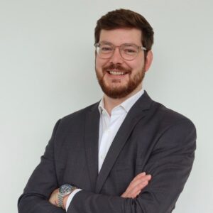 Porträt von Julius Renn, Senior Key Account Manager bei SQUEAKER. Er trägt ein weißes Hemd und ein dunkles Sakko, steht mit verschränkten Armen vor einer hellen Wand und lächelt in die Kamera.