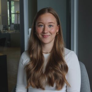 Porträt von Luisa Steffens, Werkstudentin für Eventmanagement & Customer Success bei SQUEAKER. Sie trägt ein weißes Oberteil und goldfarbene Ohrringe, sitzt lächelnd in einem modernen Büro mit Glaswänden und blickt in die Kamera.