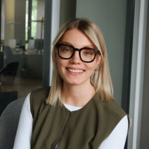 Porträt von Pia Klocke, Talent Acquisition & HR-Managerin bei SQUEAKER. Sie trägt eine dunkle Brille, ein olivgrünes Oberteil über einem weißen Shirt und sitzt lächelnd in einem modernen Büro mit Glaswänden.