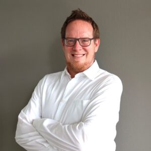 Porträt von Daniel Krewet, Head of Sales & Key Account Management bei SQUEAKER. Er trägt ein weißes Hemd und eine Brille, steht mit verschränkten Armen vor einer grauen Wand und lächelt in die Kamera.