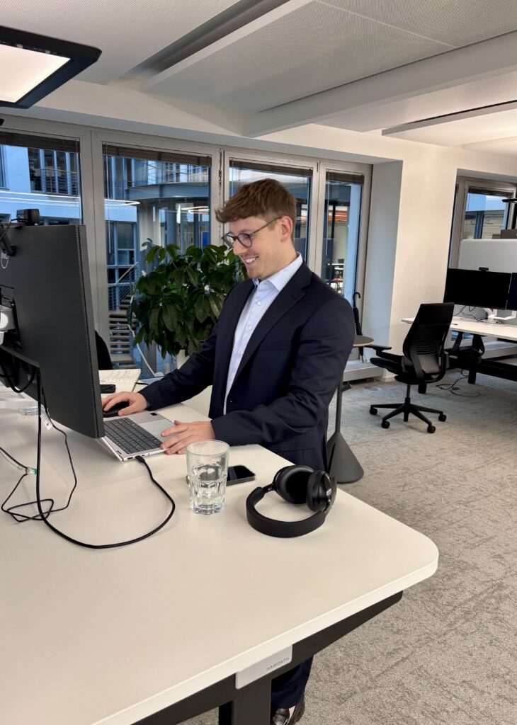 Niklas von Horvath steht in einem Büro an einem Schreibtisch mit Laptop und Monitor; auf dem Tisch liegen ein Glas Wasser und Kopfhörer.