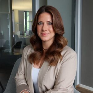 Jessica Schilles sitzt in einem Büro vor einer Glaswand und blickt in die Kamera; sie trägt einen beigefarbenen Blazer und ein weißes Oberteil.