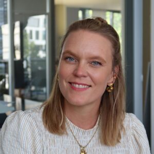 B2B final 78 Anica Iliu mit schulterlangem blondem Haar und gestreifter Bluse vor einer Fensterfront in einem Innenraum.