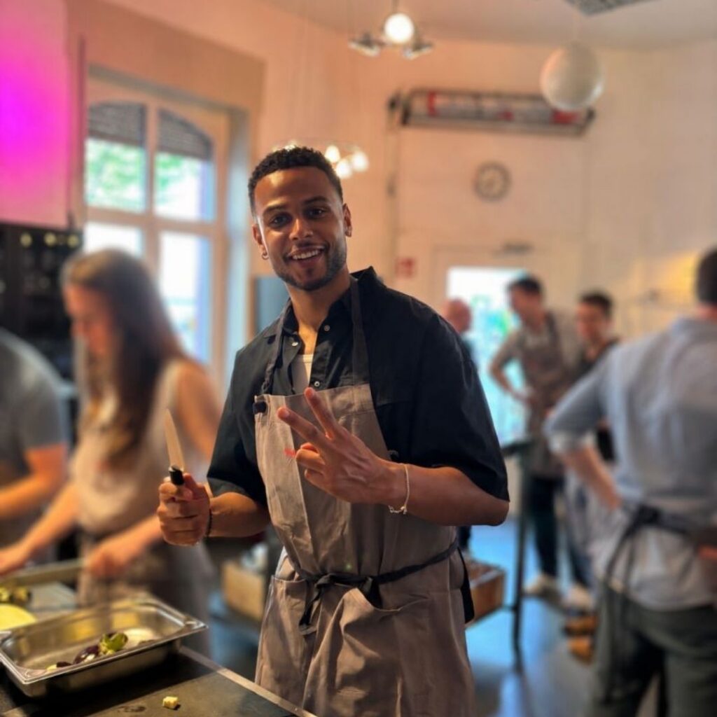 Ein Mann in schwarzem Hemd und heller Schürze steht in einer Küche und hält ein Küchenmesser in der Hand; im Hintergrund sind weitere Personen zu sehen.