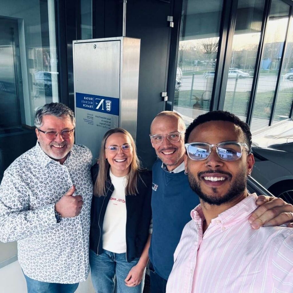 Vier Personen stehen für ein gemeinsames Foto in einem Eingangsbereich; die Person im Vordergrund trägt ein rosafarbenes Hemd und eine Sonnenbrille.