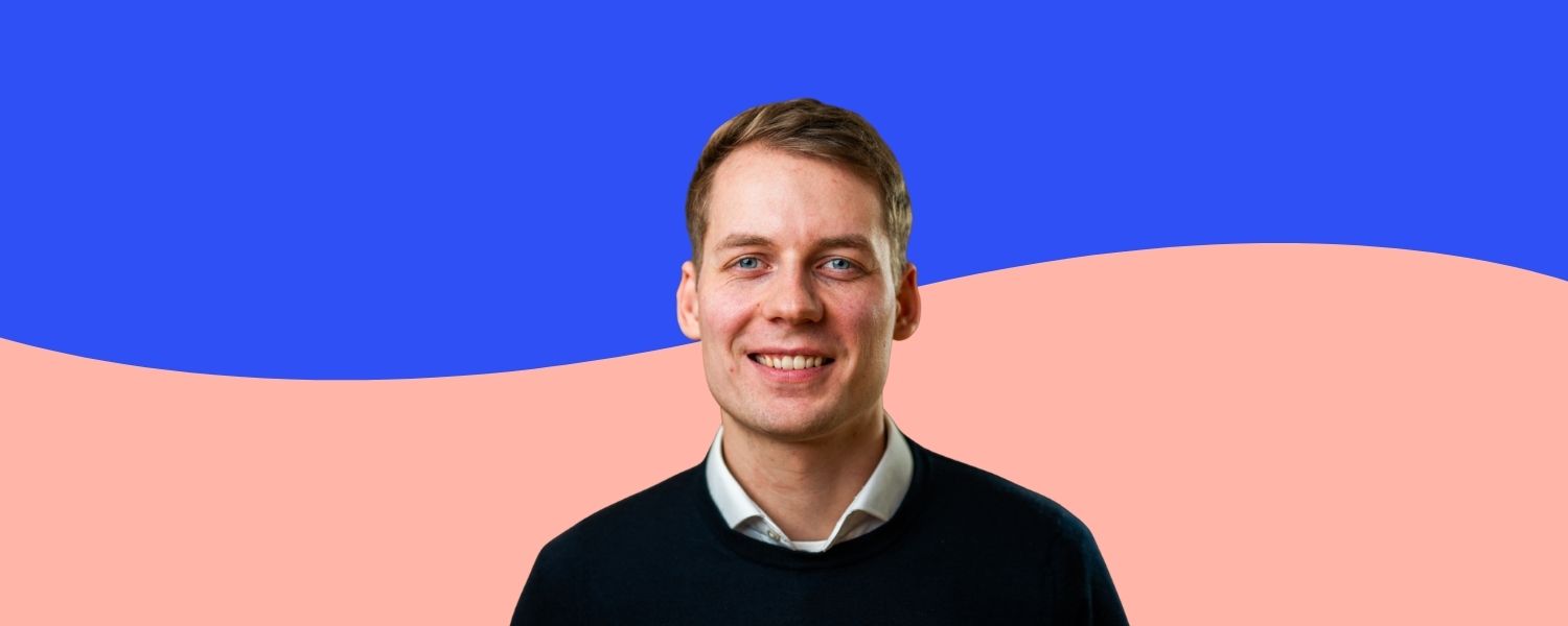 Jasper steht vor einem zweifarbigen Hintergrund in Rosa und Blau und trägt ein schwarzes Oberteil über einem weißen Hemd.