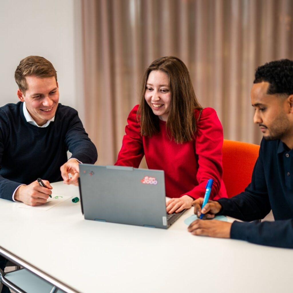 Jasper und zwei weitere Personen sitzen an einem Tisch mit einem Laptop und schreiben mit Stiften auf Papier.