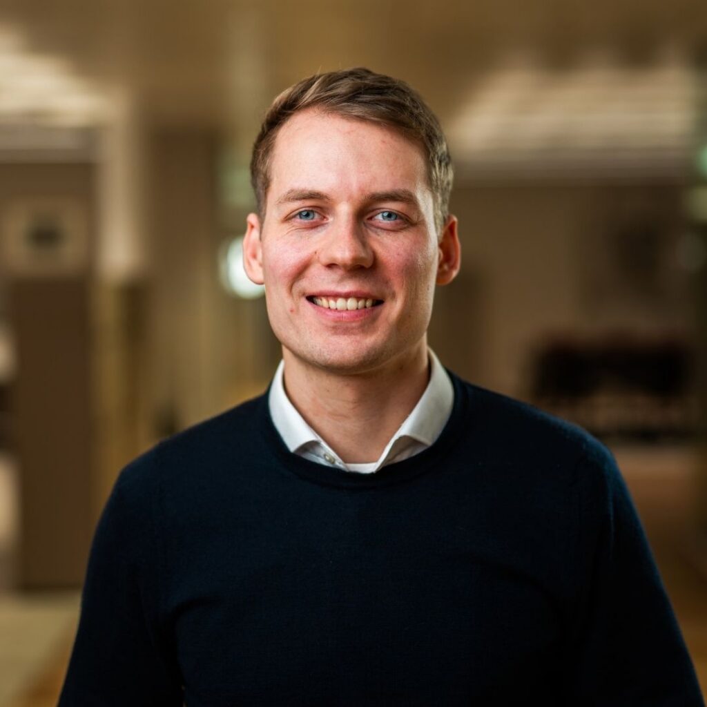 Jasper steht in einem Innenraum und trägt ein dunkles Oberteil über einem weißen Hemd.