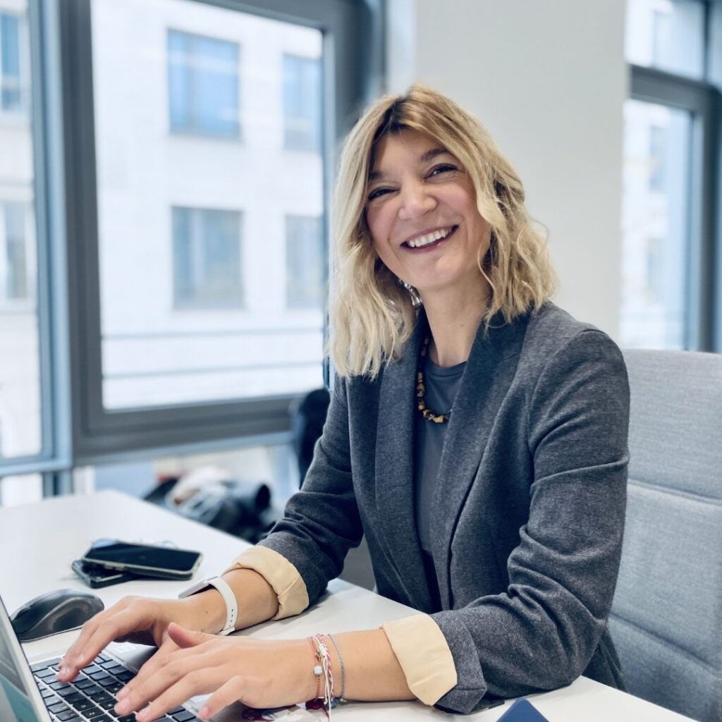 Eine Frau mit blondem Haar sitzt an einem Schreibtisch vor großen Fenstern und tippt auf einem Laptop.