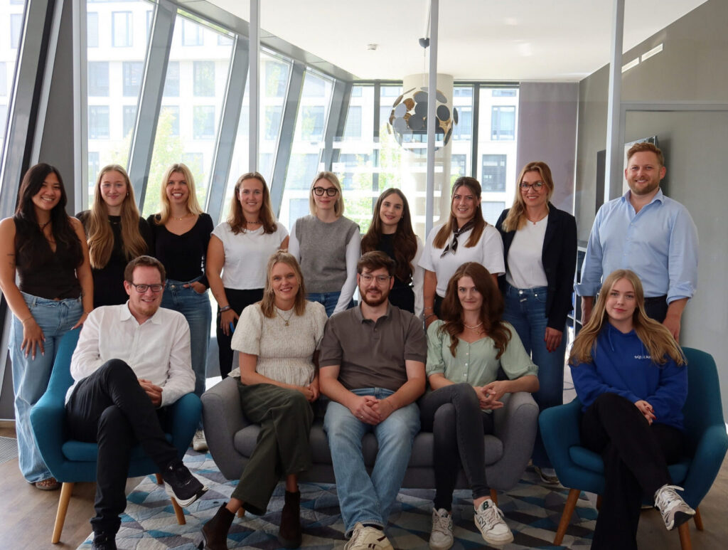 14 Personen des SQUEAKER-Teams sitzen und stehen für ein Gruppenfoto in einem hellen Büro mit großen Fenstern.
