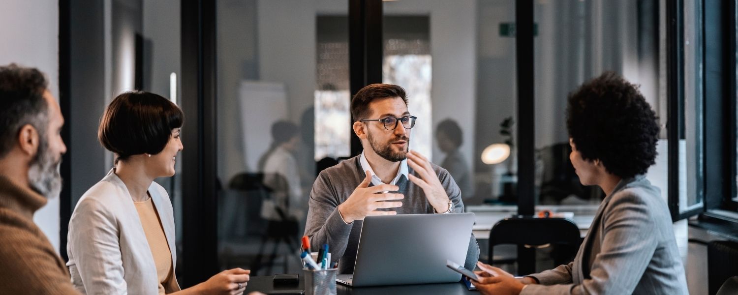 Vier Personen sitzen an einem Tisch in einem Büro; ein Mann gestikuliert hinter einem geöffneten Laptop, während die anderen zu ihm schauen.