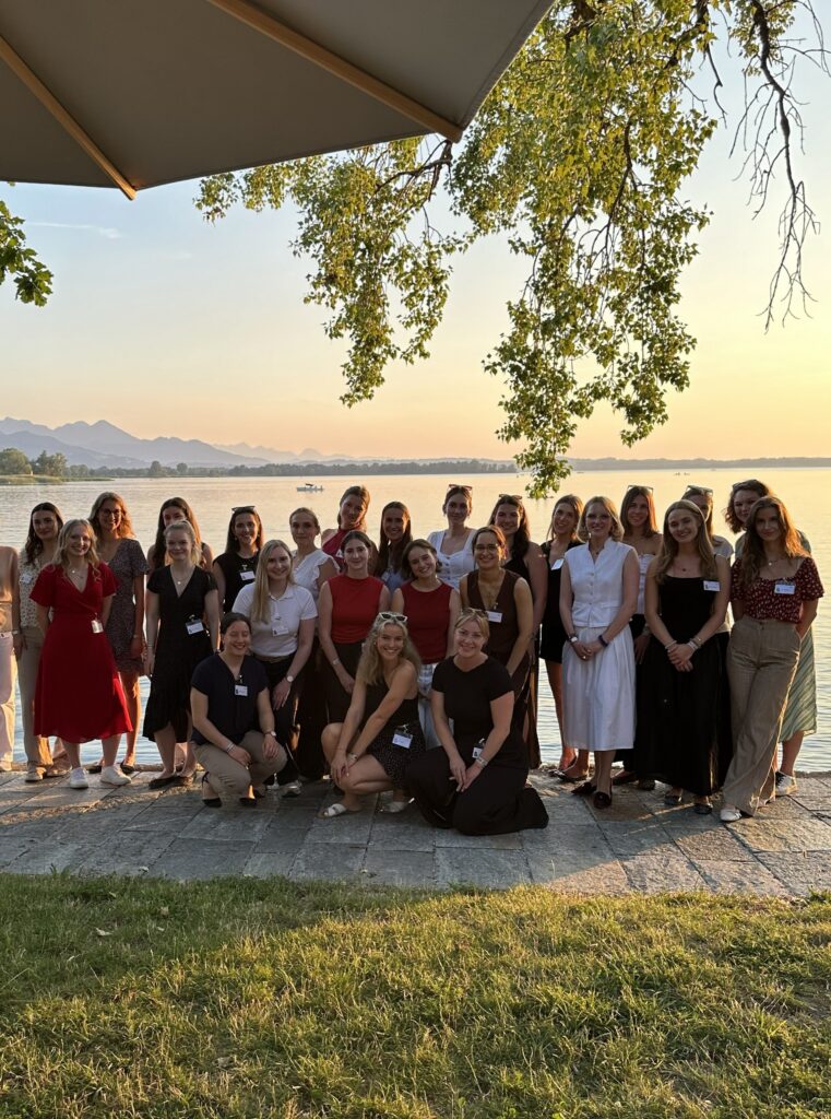 Eine Gruppe von Frauen posiert am Ufer eines Sees unter einem Baum; im Hintergrund sind das Wasser, Berge und der Abendhimmel zu sehen.