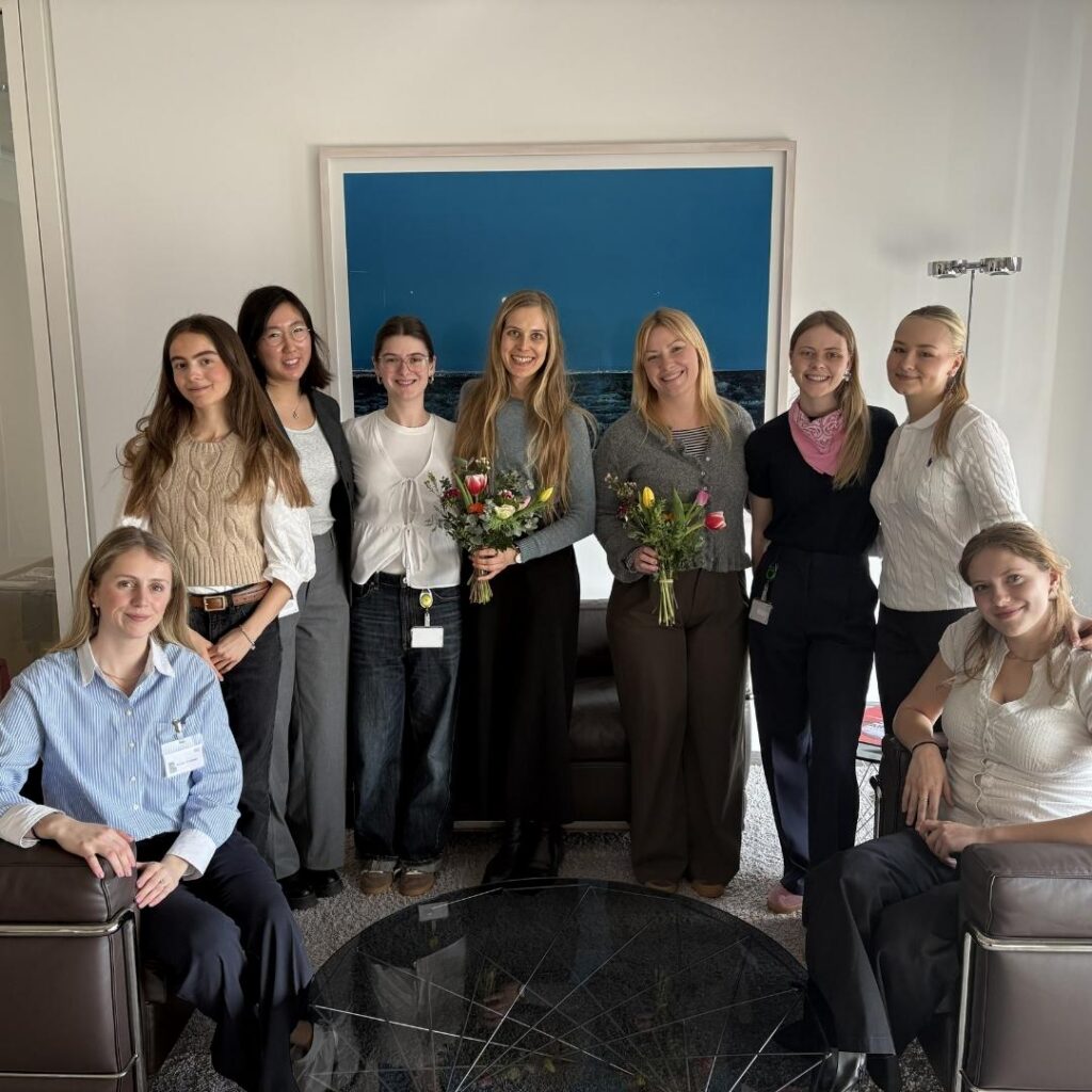 Mehrere Frauen posieren in einem Raum; zwei halten Blumensträuße, und im Hintergrund hängt ein gerahmtes Bild.